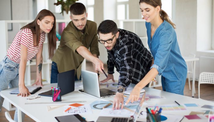 Young creative people with laptop and sketches discussing new project together. Group of guys and gals working in modern cozy office